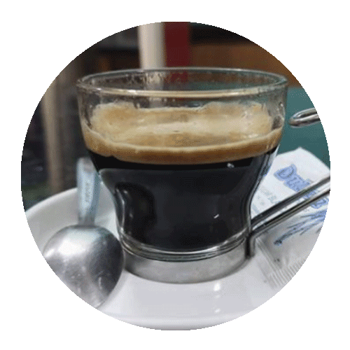 Taza de café negro servido en un vaso transparente, con espuma en la parte superior, acompañada de una cuchara y un sobre de azúcar.